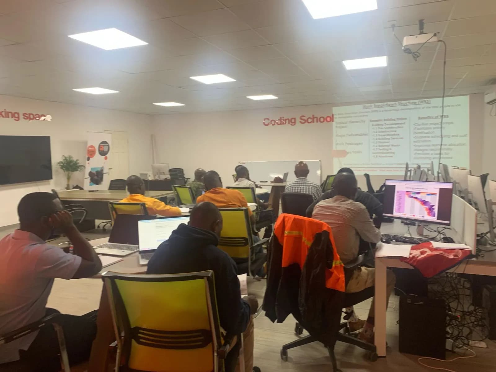 A class of adult students at tables with laptops, facing a projected slide on the work breakdown structure