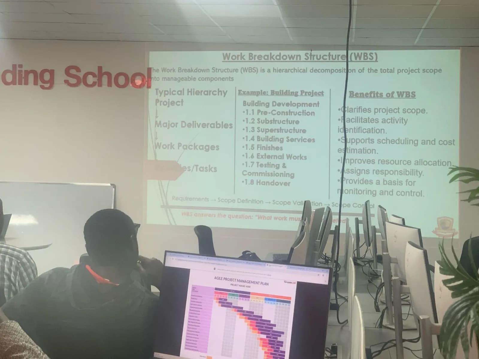 A projected slide explaining the work breakdown structure of a building project, with an agile project plan on a monitor in front